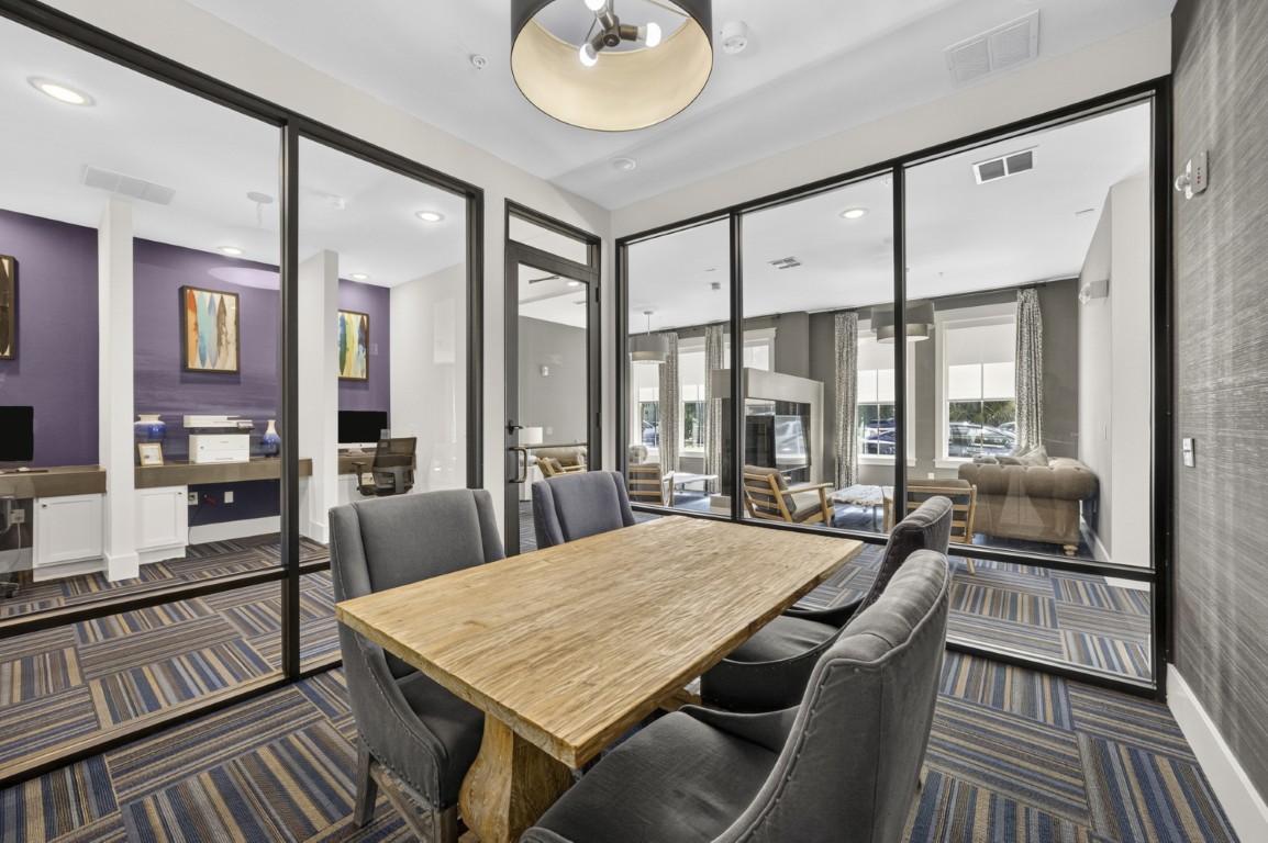 Novus Conference room with a wooden table, gray chairs, and glass walls overlooking a lounge area with couches and computers.