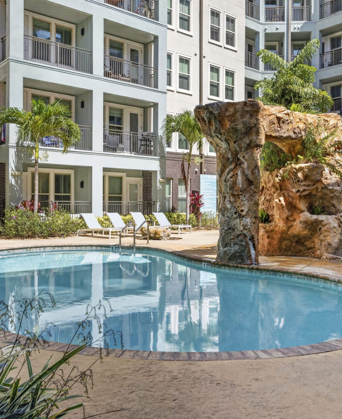 Novus Outdoor apartment pool with lounge chairs, palm trees, and a rock waterfall feature, surrounded by multi-story buildings.